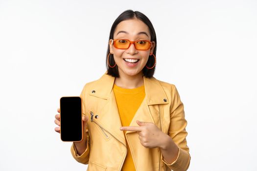 Portrait of stylish korean girl in sunglasses, smiling, pointing finger at smartphone screen, showing mobile phone application, standing over white background.