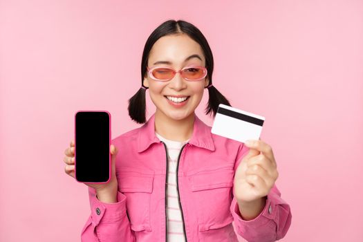 Image of smiling korean woman showing credit card and mobile phone screen, smartphone application interface, paying online, shopping contactless, standing over pink background.