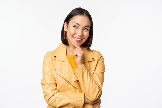 Portrait of stylish korean woman in yellow jacket, smiling thoughtful, thinking and looking up at logo or advertisement, standing over white background.