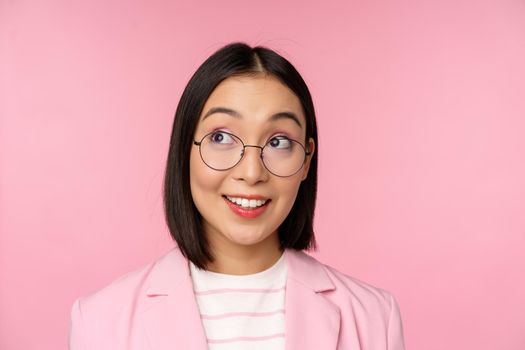 Enthusiastic saleswoman, asian businesswoman in glasses, looking intrigued at upper left corner logo, company advertisement, standing over pink background.