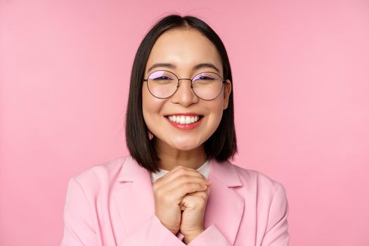 Close up portrait of smiling happy businesswoman in glasses, clench hands together thankful, excited of smth, begging or say please, standing over pink background.