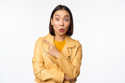 Portrait of beautiful asian woman express intereest and excitement about advertisement, store discount, pointing finger left, looking amazed, standing over white background.