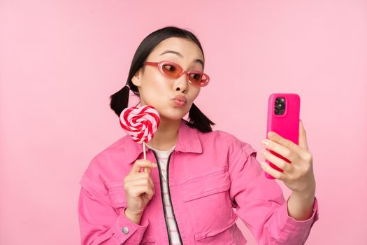 Portrait of stylish, happy asian girl taking selfie with candy, lolipop sweets and smiling, taking photo with mobile app, standing over pink background.