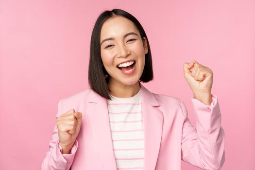 Enthusiastic saleswoman, asian corporate woman say yes, achieve goal and celebrating, triumphing, looking with rejoice and smiling, standing over pink background.