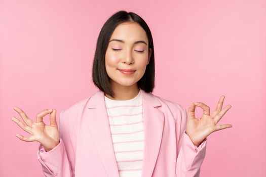 Keep calm. Smiling young asian woman meditating, practice yoga, mindfulness and relaxation at work, breath, inhale air with pleased face, standing over pink background.