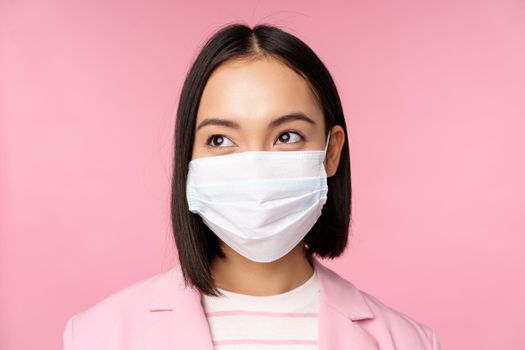 Close up portrait of japanese corporate woman in medical face mask from covid-19, looking left at logo, sale promo, standing over pink background.