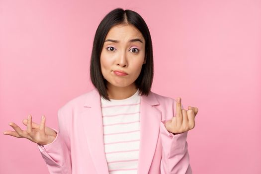 Portrait of young asian business woman, saleswoman shrugging shoulders and looking confused, clueless of smth, standing over pink background.