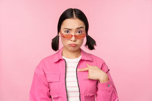 Portrait of asian girl looks confused and points at herself, perplexed face, stares with disbelief at camera, stands over pink background.
