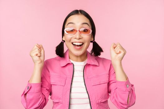Portrait of excited japanese girl in sunglasses, celebrating, achieve goal, gasping amazed and smiling, standing over pink background.