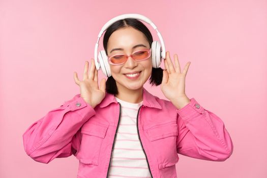 Dancing stylish asian girl listening music in headphones, posing against pink background.