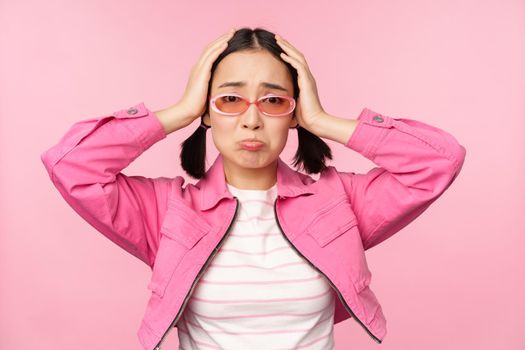 Portrait of troubled asian woman, korean girl holds hands on head and look in panic, standing distressed and shocked against pink background.