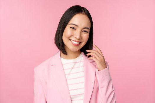 Young professionals. Smiling asian businesswoman, saleswoman in suit looking confident at camera, posing against pink background.