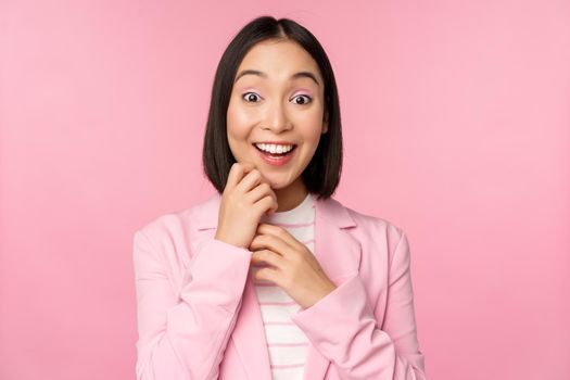 Happy young office lady, korean business woman wearing suit, looking surprised at camera, posing against studio background in pink.