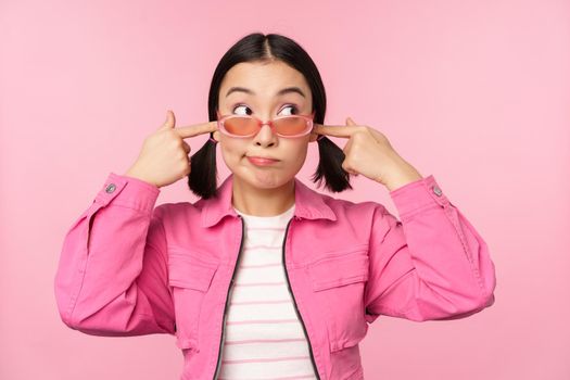 Portrait of silly japanese girl shuts ears, looks aside, doesnt listen, cant hear you gesture, standing over pink background, ignoring you.