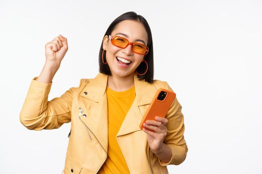 Enthusiastic korean girl in sunglasses, holding smartphone, celebrating and dancing, laughing happy with mobile phone, standing over white background.