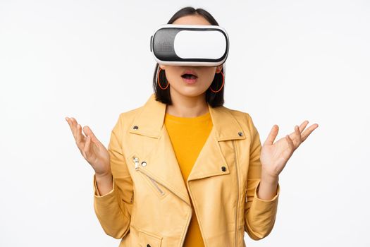 Young asian girl wearing VR glasses, looking amazed, using virtual reality headset, posing against white background.
