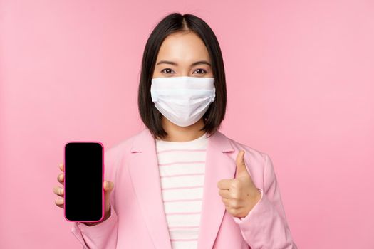 Portrait of asian corporate woman in medical face mask covid-19, showing smartphone screen and thumb up sign, pink background.
