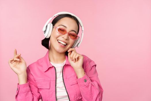 Dancing stylish asian girl listening music in headphones, posing against pink background.