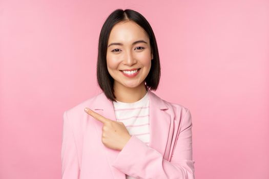 Portrait of asian business woman, saleswoman in suit pointing finger left, showing banner advertisement, smiling and looking professional, pink background.