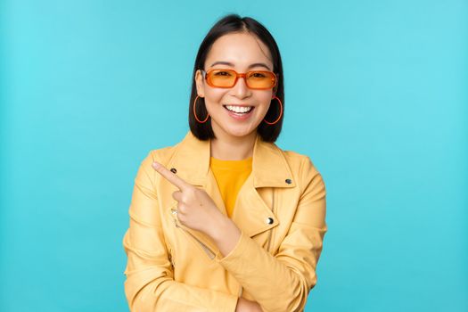 Smiling asian woman in sunglasses, pointing finger left, showing banner, logo or advertisement, standing over blue background. Copy space