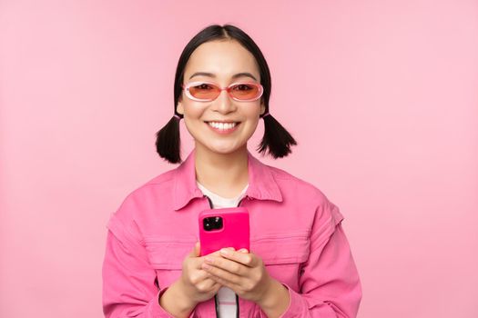 Portrait of korean girl in stylish sunglasses holding mobile phone, using smartphone app, standing over pink background. Copy space