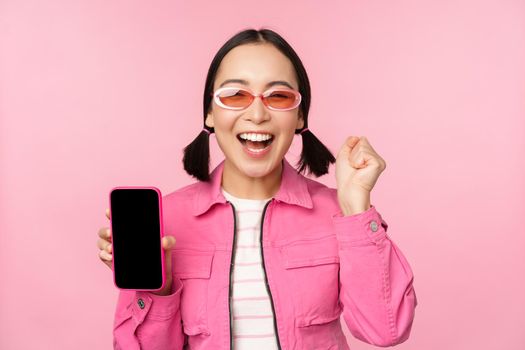 Excited asian girl laughs and smiles, shows mobile phone screen, smartphone application, standing over pink background.