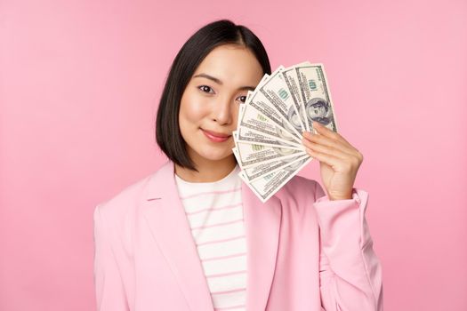 Microcredit, investment and business people concept. Young asian businesswoman, corporate lady showing money, cash dollars, smiling pleased, pink background.