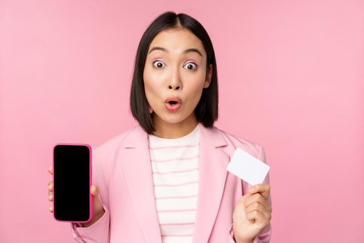 Enthusiastic asian businesswoman showing mobile phone screen and credit card, looking amazed at camera, standing over pink background.