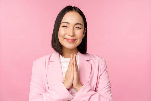 Thank you. Smiling asian saleswoman, corporate lady in suit thanking, holding hands in bag, gratitude gesture, standing over pink background.