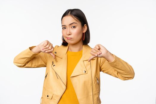 Image of fully disappointed asian woman showing thumbs down, shaking head displeased, dislike smth, standing over white background.