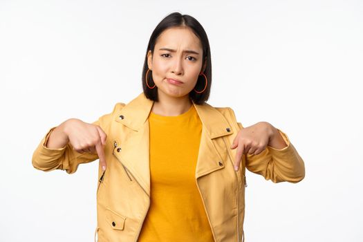 Portrait of disappointed asian woman pointing fingers down, grimacing and showing smth bad, standing over white background. Copy space
