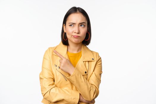 Asian girl looking skeptical, pointing finger left at smth strange, staring doubtful and displeased at product banner, standing against white background.
