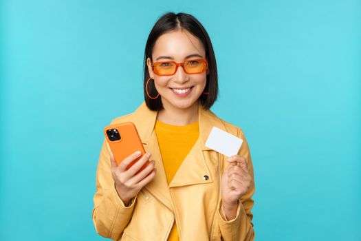 Online shopping. Stylish young asian woman in sunglasses, showing credit card and using smartphone, paying in internet, making purchase, standing over blue background.