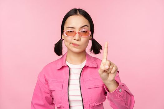 Image of serious, stylish asian girl in sunglasses, showing stop, prohibit gesture, taboo sign, saying no, standing over pink background.