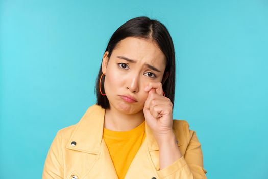 Close up of sad asian girl crying, wiping tears and sulking, looking upset and gloomy, posing against blue background. Copy space