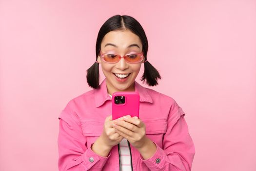 Portrait of asian girl in sunglasses using smartphone. Woman looking at mobile phone, browsing in app, standing over pink background.