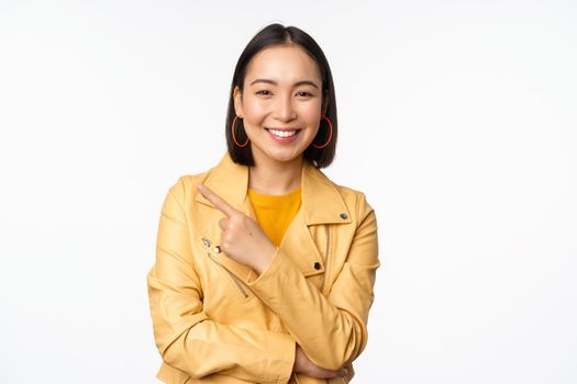 Portrait of smiling asian girl pointing finger left, showing logo or copy space, advertisement on empty side, standing over white background.