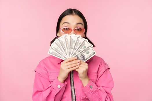 Beautiful korean woman in sunglasses, showing dollars, money cash, smiling pleased, concept of fast loans, microcredit and payment, standing over pink background.