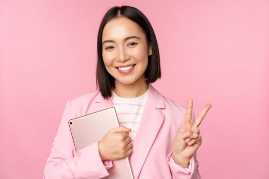Portrait of asian businesswoman in suit, standing with digital tablet, showing peace, v-sign and smiling, pink background.
