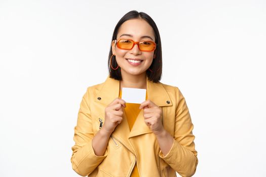 Portrait of smiling korean female model in sunglasses, showing credit card, standing over white background.