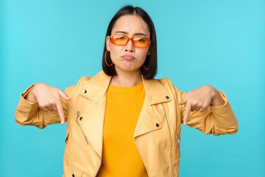 Sad and gloomy chinese girl in sunglasses, points fingers down and grimaces, looks miserable and disappointed, emotion of regret, standing over blue background.