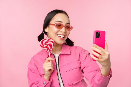 Portrait of stylish, happy asian girl taking selfie with candy, lolipop sweets and smiling, taking photo with mobile app, standing over pink background.