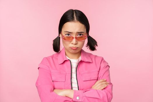Portrait of sad and gloomy asian girl sulking from disappointment, standing upset against pink studio background. Copy space