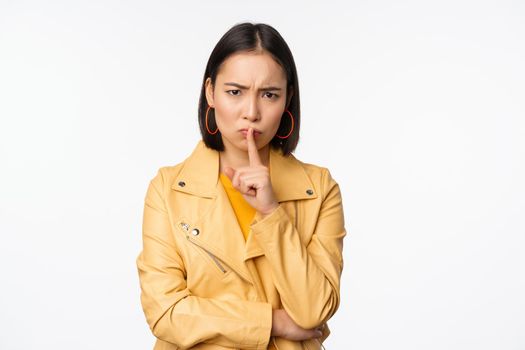 Portrait of angry korean girl shushing, woman frowning and tell to be quiet, showing hush, shh gesture, press finger to lips, taboo sign, standing over white background.