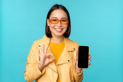 Smiling korean woman showing mobile phone app interface, smartphone application, recommending on cellphone, standing over blue background.