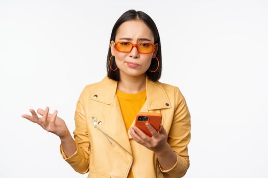 Portrait of confused asian girl in sunglasses, holding smartphone, using mobile phone and looking puzzled, standing clueless over white background.