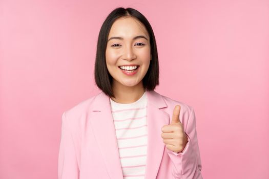 Portrait of asian businesswoman smiling satisfied, showing thumbs up, praise, like and approve, standing in suit over pink background.