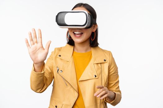 Happy asian girl in VR headset, saying hello in virtual reality chat, using special glasses, standing over white background, waving hand, saying hello.