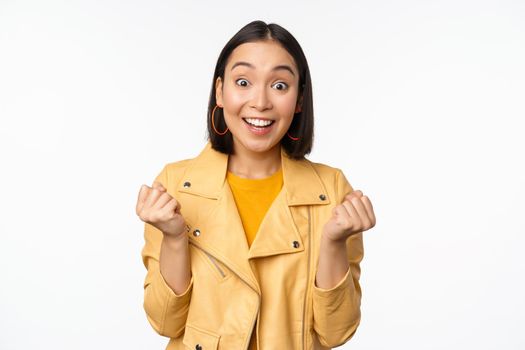 Surprised and happy asian girl, looking with happy face of rejoice, winning, celebrating victory and triumphing, standing over white background.