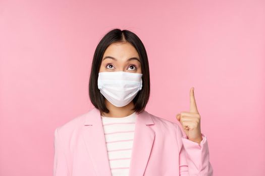 Close up portrait of businesswoman, asian lady in office suit and medical face mask, looking and pointing up, showing company logo or banner on top, pink background.